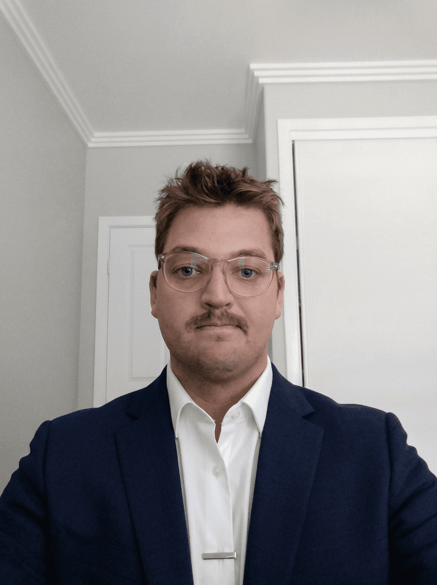 Man with clear glasses and a mustache wearing a navy blue blazer and white shirt.