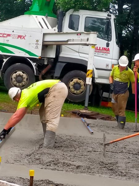 Construction workers leveling wet concrete with tools in front of a white concrete pump truck.